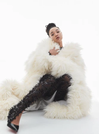 Woman seated on a white seamless backdrop wearing an oversized shaggy white coat, embellished black tights, and black patent high heels.