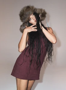 Fashion model in a brown faux fur trapper hat and burgundy sleeveless mini dress posing against a pale grey seamless studio backdrop.