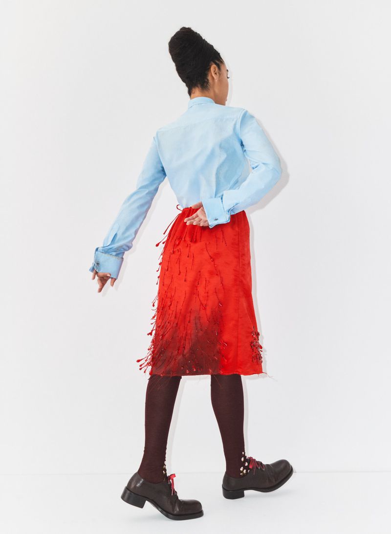 Model seen from behind wearing a light blue dress shirt, bright red fringed skirt, dark tights, and lace-up shoes against a white background.