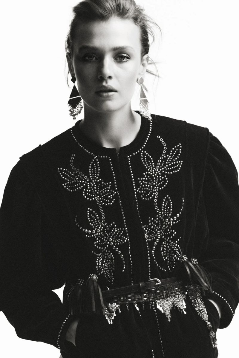 Monochromatic studio shot of a woman with fair skin wearing a studded black velvet jacket, statement earrings, and a decorative belt.