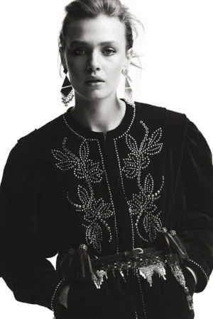 Monochromatic studio shot of a woman with fair skin wearing a studded black velvet jacket, statement earrings, and a decorative belt.