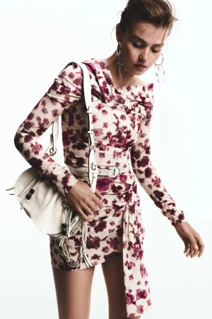 Model Ella Mccutcheon poses in a floral print mini dress with an off-shoulder neckline and white shoulder bag against a white backdrop.