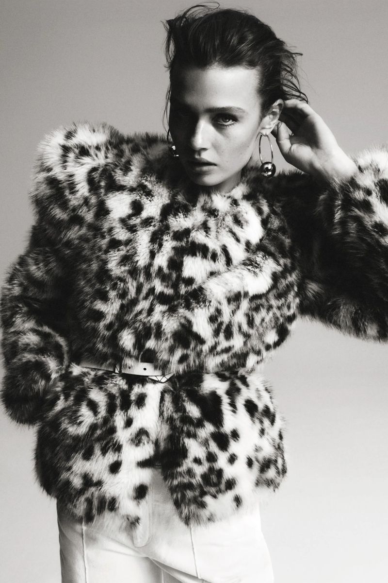 A woman with dark hair poses in a fuzzy, spotted jacket with a belt and white pants against a light gray studio backdrop.