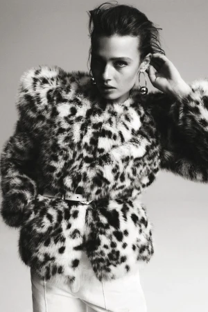 A woman with dark hair poses in a fuzzy, spotted jacket with a belt and white pants against a light gray studio backdrop.