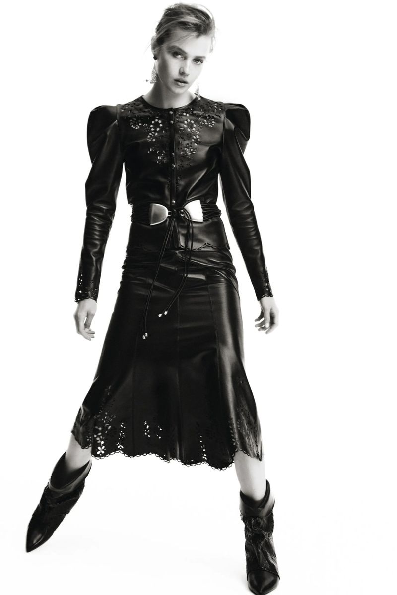 Monochrome fashion shot of a woman in a leather skirt, top, belt and cowboy boots, against a white backdrop in a studio setting.
