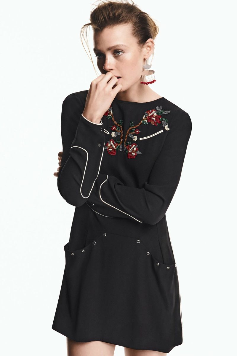 A fair-skinned young woman poses indoors wearing a black dress with floral embroidery, set against a simple, bright white backdrop.