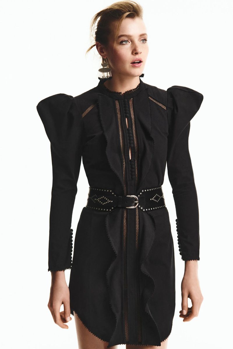 Woman in a black dress with puffed sleeves and a studded belt poses against a white background in a fashion studio setting.