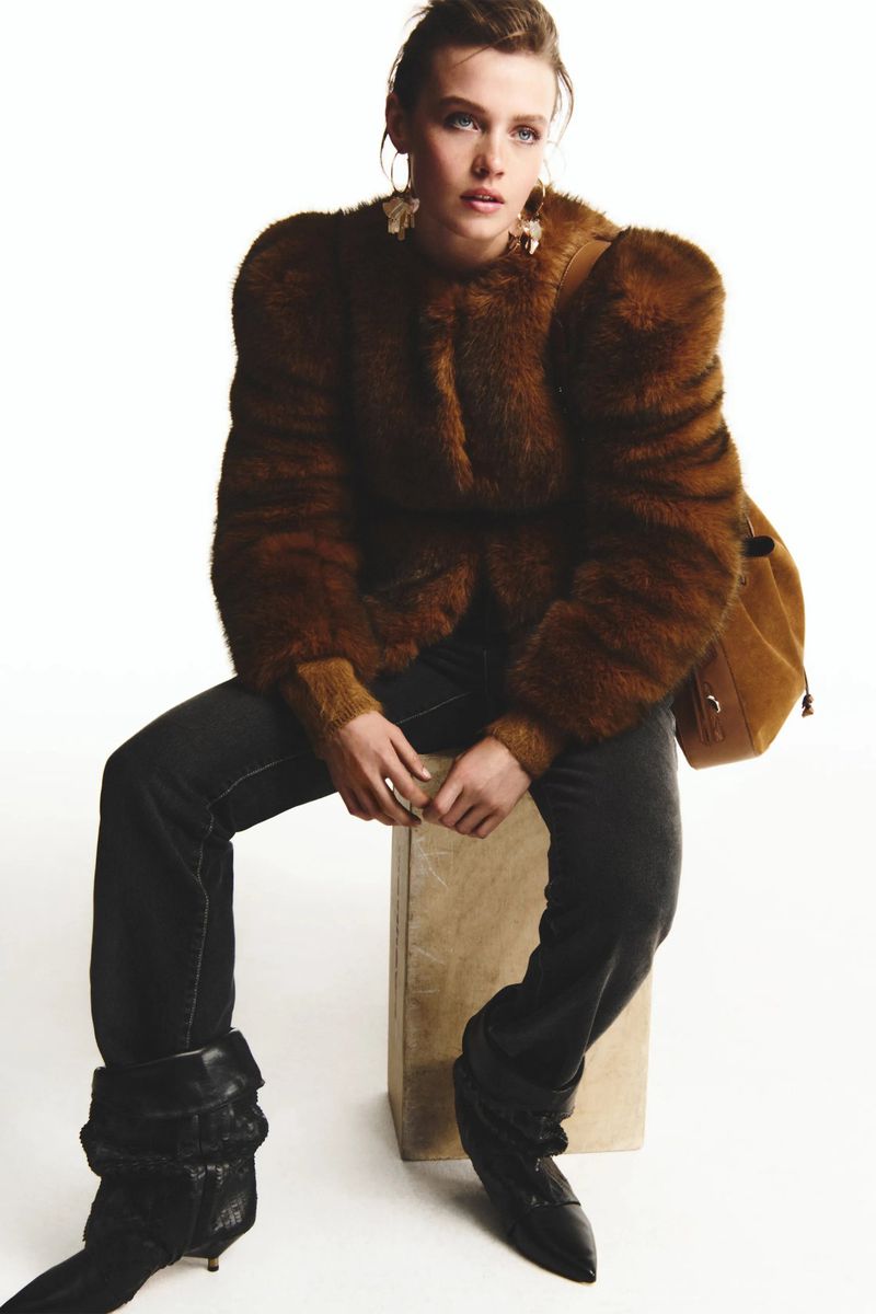 A woman poses in a brown faux fur jacket, dark denim jeans, and black leather boots while sitting on a tan cube against a white backdrop.
