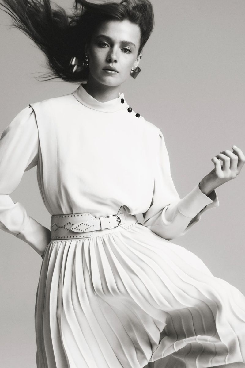 Monochromatic portrait of woman in white long-sleeve dress with pleated skirt and statement earrings against a grey background.