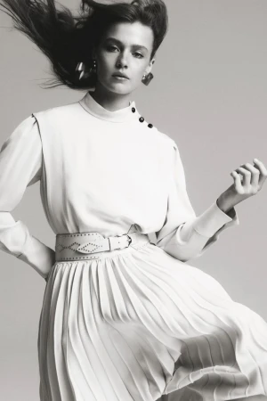 Model Ella McCutcheon wearing a long-sleeved high-neck pleated dress with a wide studded belt against a studio backdrop.