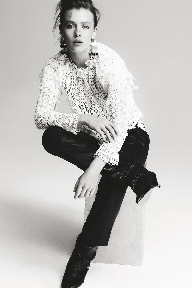 A young woman in a white lace top and dark pants poses against a neutral background in a studio shot, looking directly at the camera.
