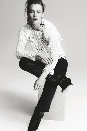 A young woman in a white lace top and dark pants poses against a neutral background in a studio shot, looking directly at the camera.