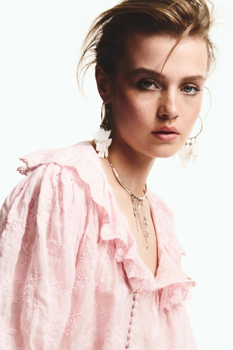 Close-up of a woman with fair skin, wearing a light pink blouse with ruffled collar and statement earrings, posing against a white background.
