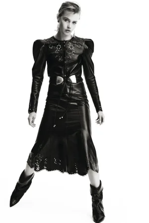 Model wearing a black leather outfit with embroidered detailing and metallic belt against a seamless white background.