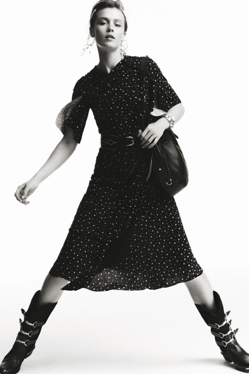 Model in polka dot dress and buckled boots holding a black leather handbag against a plain white background