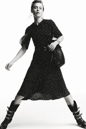Model in polka dot dress and buckled boots holding a black leather handbag against a plain white background