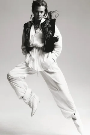 Model in motion wearing a white blouse, trousers, and embroidered black vest against a plain background