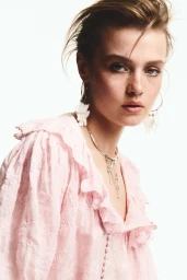 Model Ella Mccutcheon wearing a pale pink embroidered ruffle blouse and dangling earrings against a white studio background.