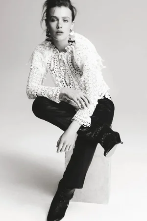 Model seated on a block wearing a patterned white lace blouse and dark trousers against a plain seamless background.