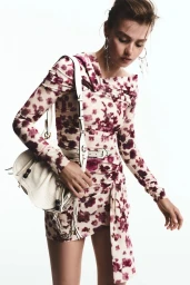 Model Ella Mccutcheon poses in a floral print mini dress with an off-shoulder neckline and white shoulder bag against a white backdrop.