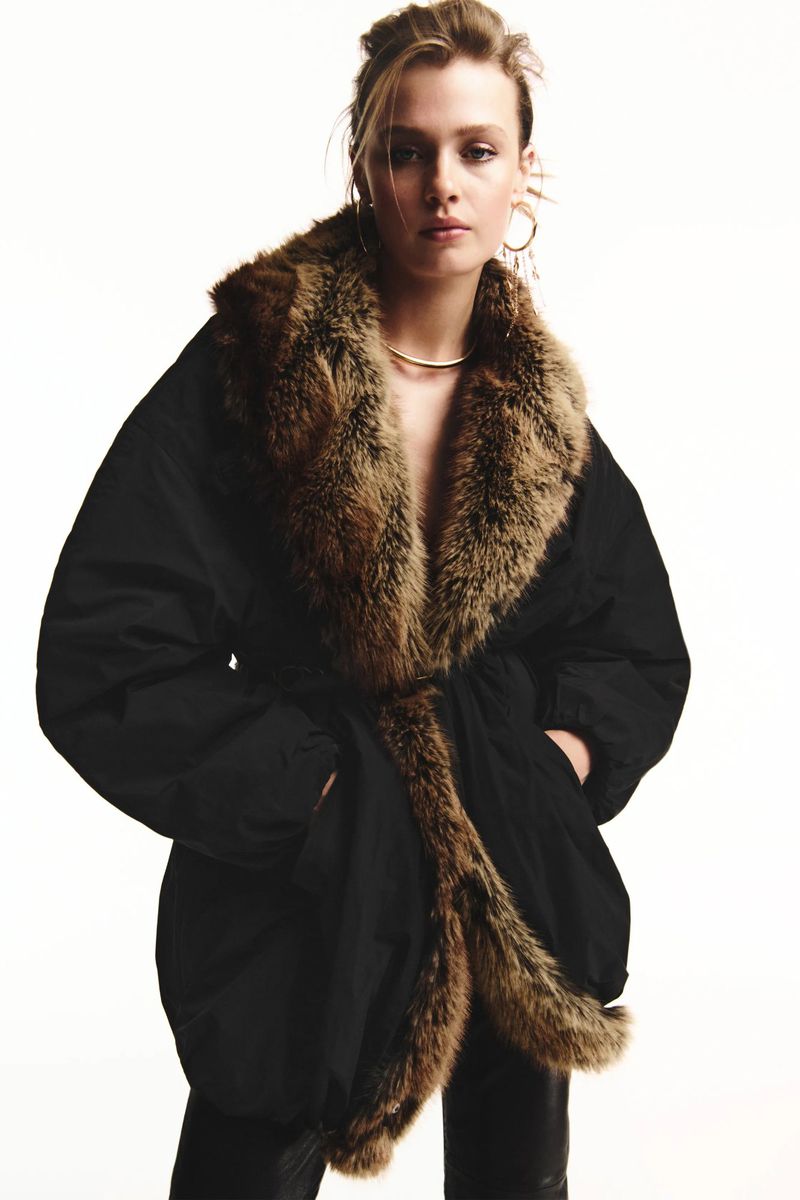 Model Ella McCutcheon wears an oversized black parka with plush brown fur trim against a bright white background.