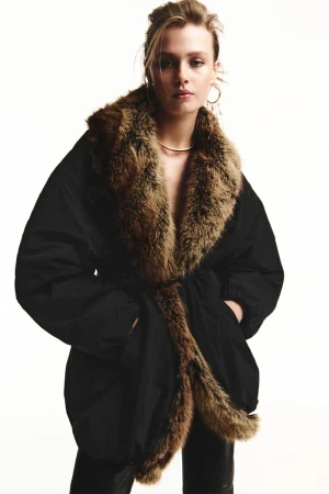 Model Ella McCutcheon wears an oversized black parka with plush brown fur trim against a bright white background.