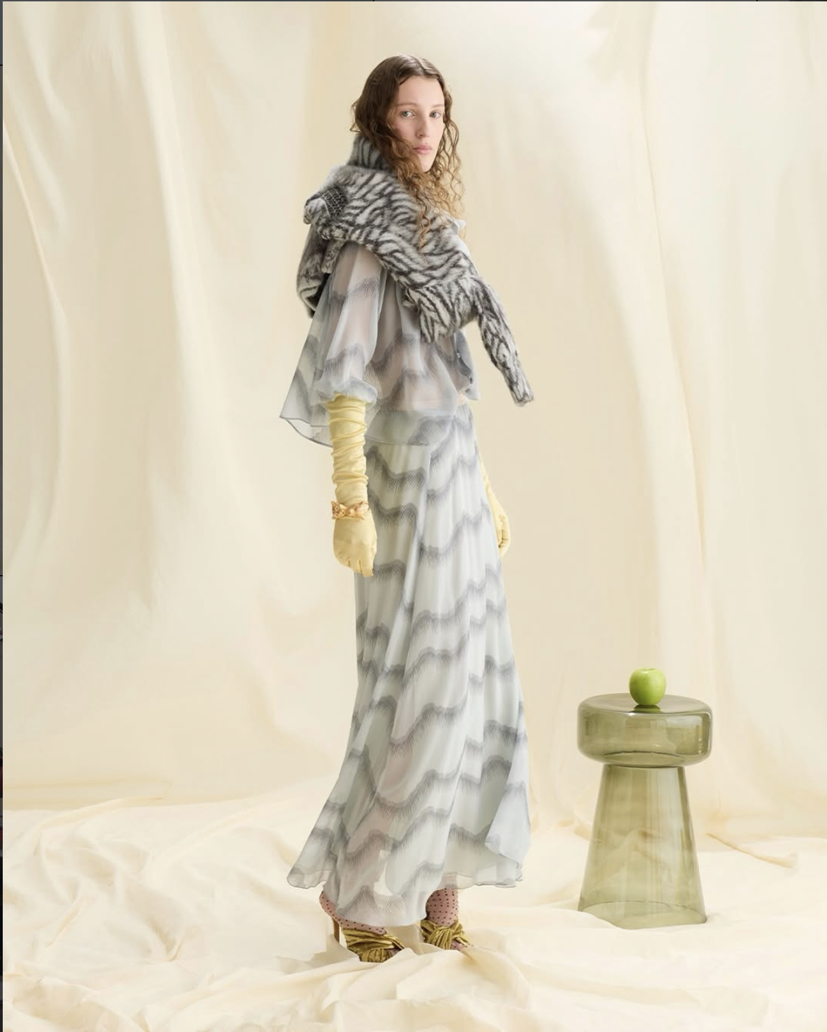 Model standing in a wavy-patterned grey maxi dress, shoulder wrap, and yellow opera gloves beside a green glass stool with an apple.