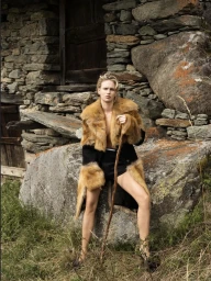 Model seated on a stone rock wearing a brown fur coat and holding a wooden stick before a rustic stone building.
