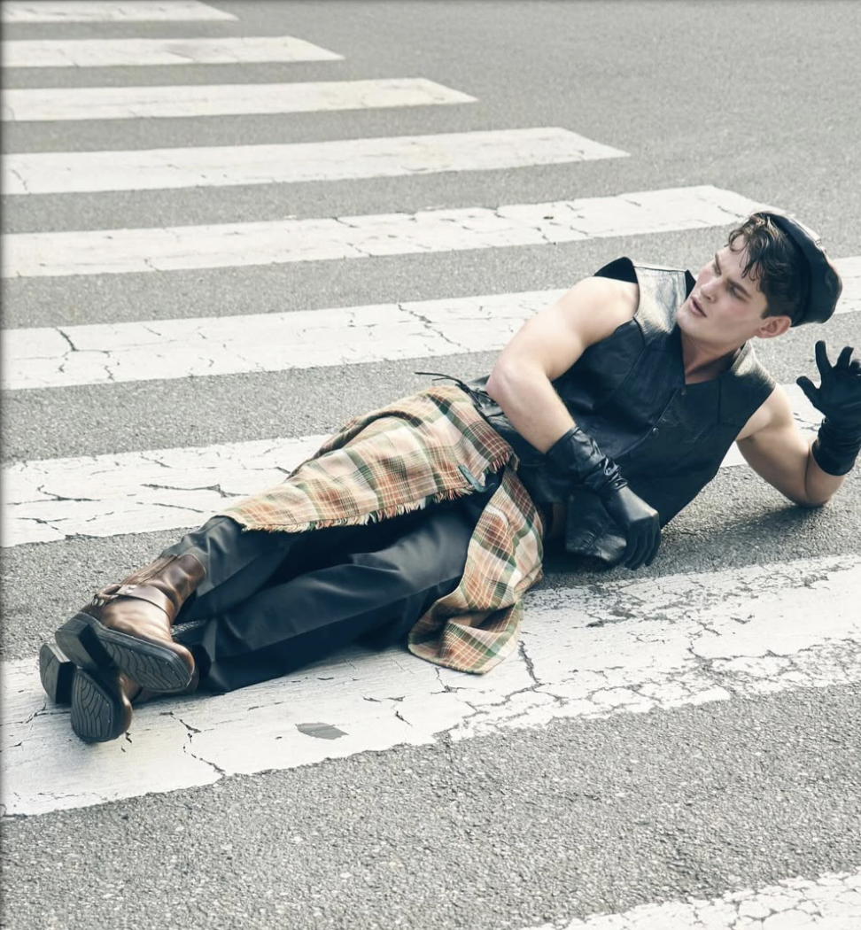 Man lying across a street crosswalk wearing a black sleeveless leather vest, gloves, plaid wrap, and brown boots.