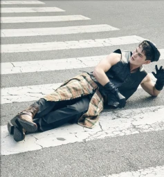 Man lying across a street crosswalk wearing a black sleeveless leather vest, gloves, plaid wrap, and brown boots.