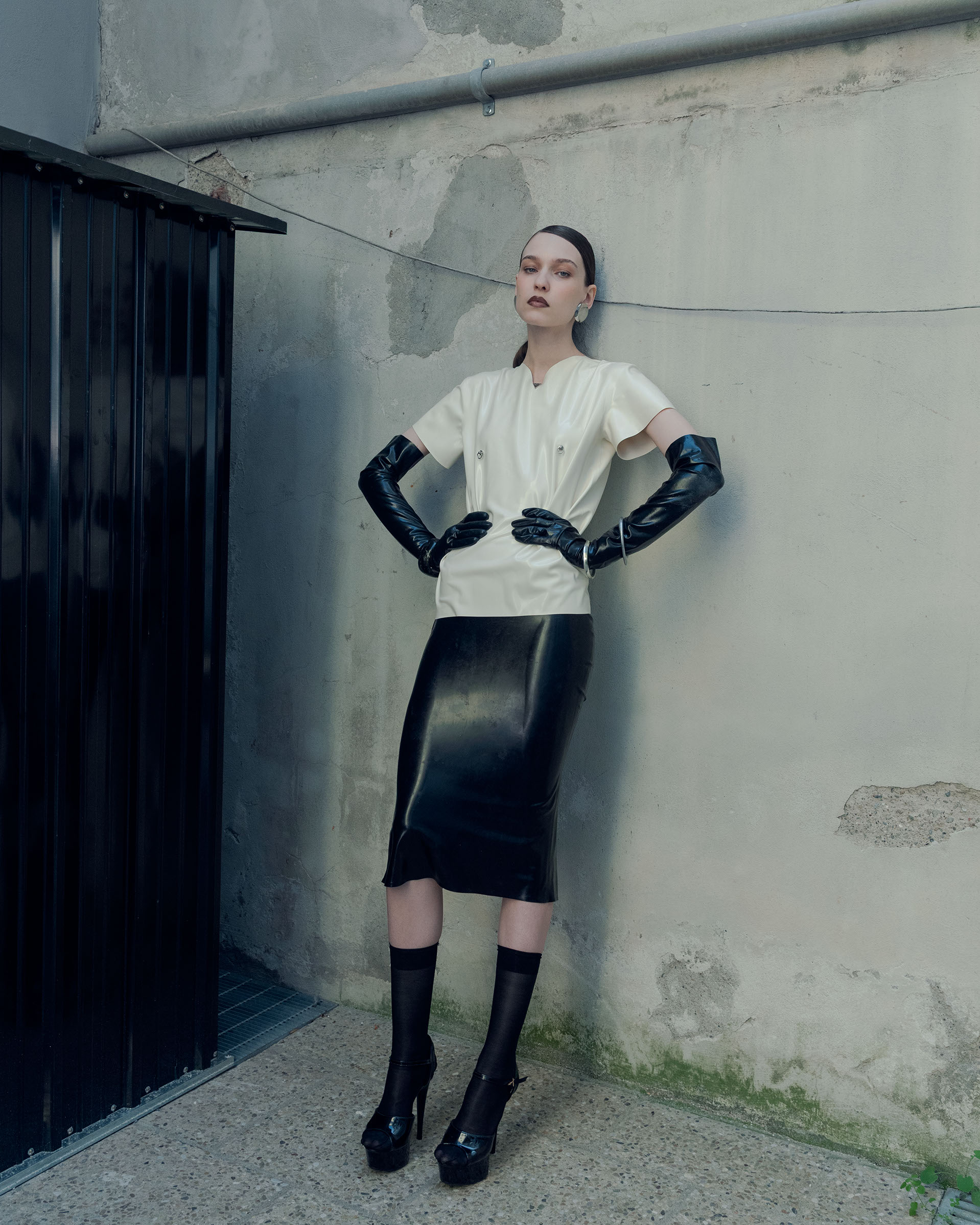 Model wearing a cream short-sleeve top, black latex midi skirt, and long black opera gloves leaning against a concrete wall.