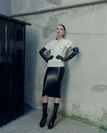 Model wearing a cream short-sleeve top, black latex midi skirt, and long black opera gloves leaning against a concrete wall.