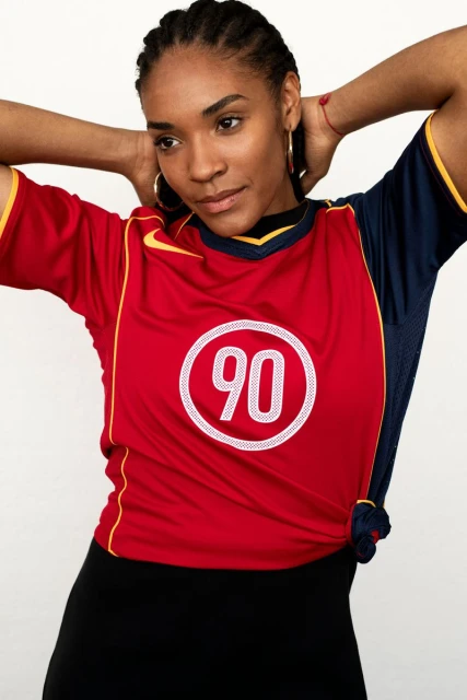 Woman in a red and navy blue sports jersey with the number 90 stands in a studio with hands behind her head.