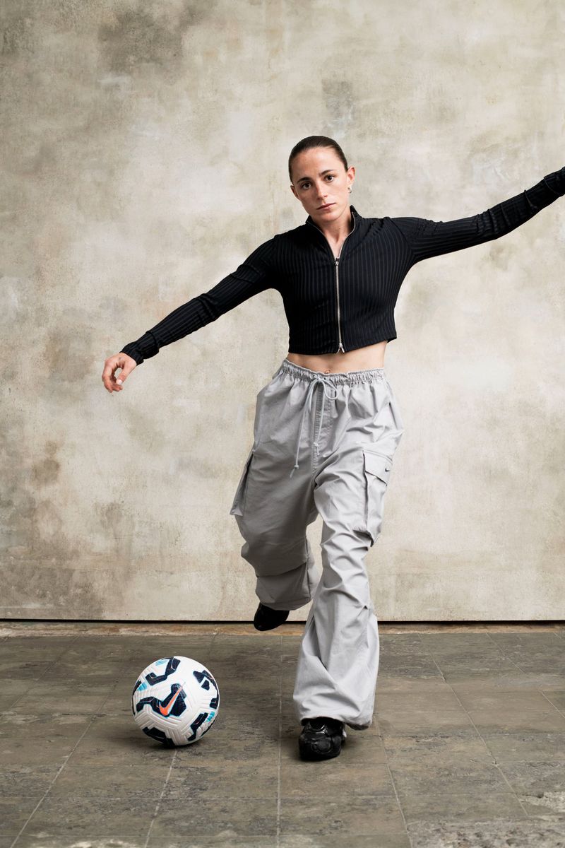 Female athlete in black top and grey pants kicks a soccer ball in a photo studio against a textured backdrop, creating a dynamic sports image.