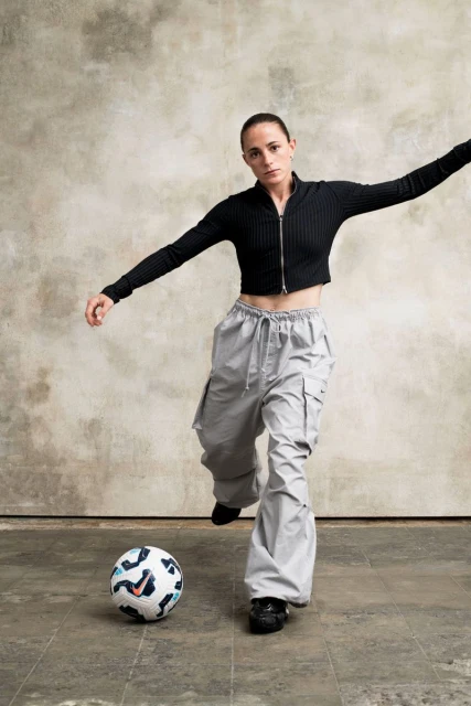 Female athlete in black top and grey pants kicks a soccer ball in a photo studio against a textured backdrop, creating a dynamic sports image.