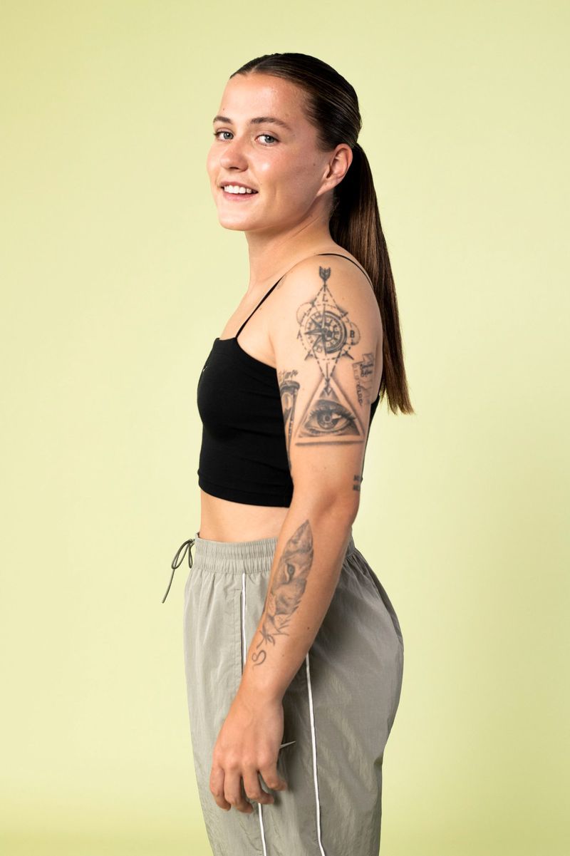 Profile shot of an adult woman with black tank top and grey sweatpants with dark brown ponytail, tattoos on arms, and light green background.