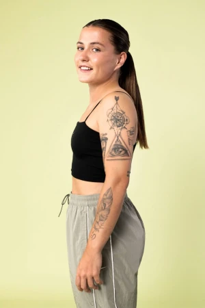 Profile shot of an adult woman with black tank top and grey sweatpants with dark brown ponytail, tattoos on arms, and light green background.