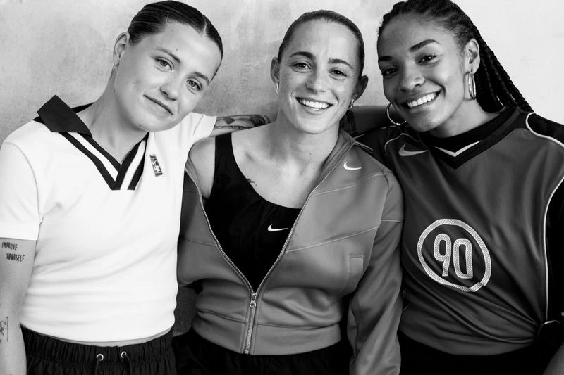 Three smiling women pose indoors. One wears a white collared shirt, another a black sports top and grey jacket, and the third a sports jersey.
