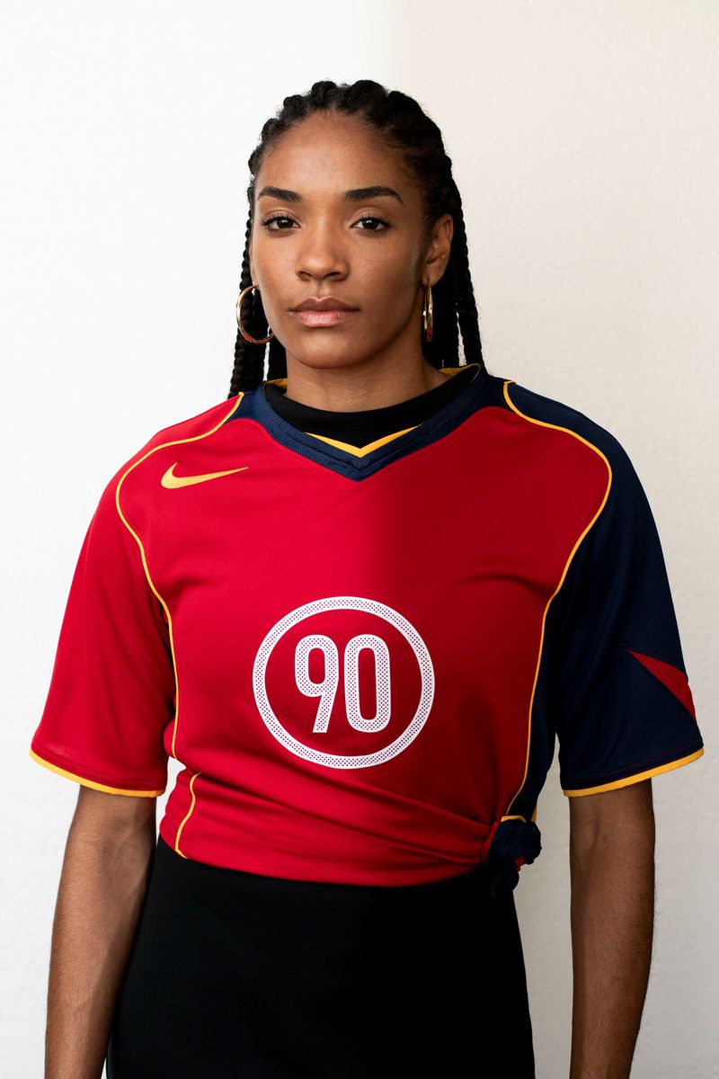 Adult woman wearing a red and blue athletic jersey with the number 90, gold hoop earrings, and a black skirt, posing indoors.