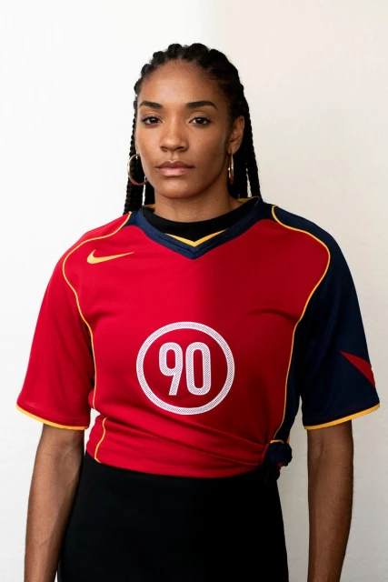Adult woman wearing a red and blue athletic jersey with the number 90, gold hoop earrings, and a black skirt, posing indoors.