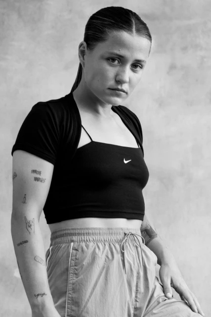 Portrait of a young adult female with tattoos, wearing a black top and grey sweatpants, posing in front of a grey background.