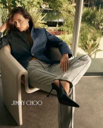 Woman in blue blazer and grey trousers seated in a modern chair wearing black slingback heels for Jimmy Choo.