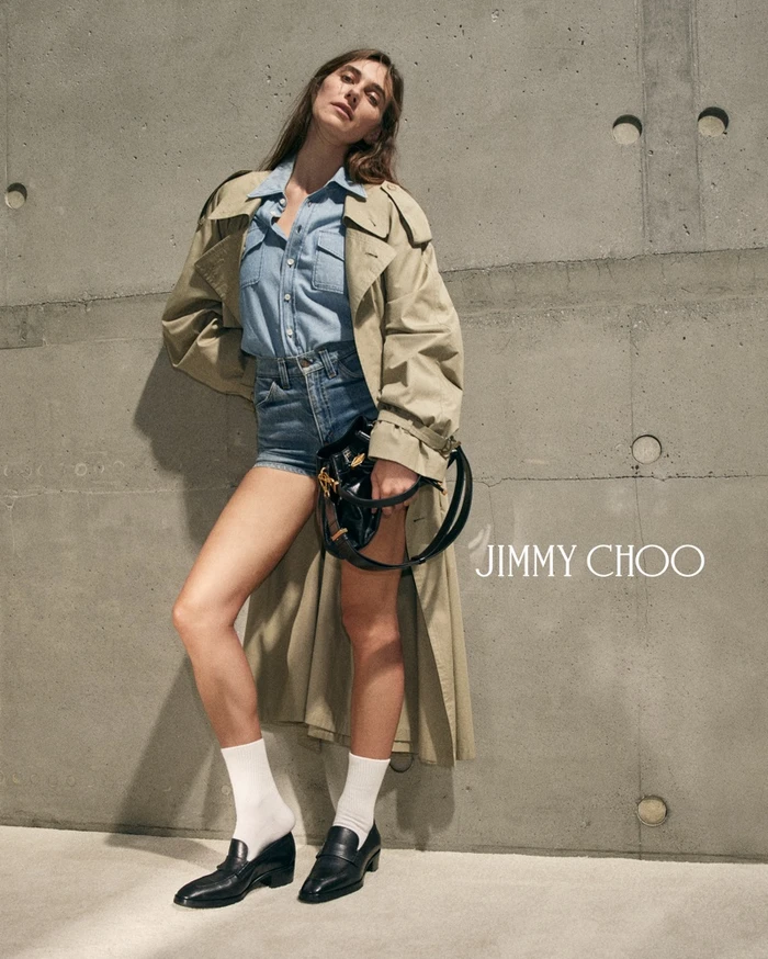 Devon Lee Carlson posing against a raw concrete wall in a khaki trench coat, blue denim shirt, shorts, white socks, and black loafers.