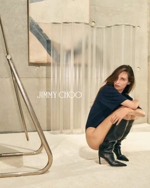 Model wearing black high-heel boots and a dark blue shirt squatting in a modern concrete room with a metal frame and artwork.