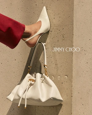 Foot in white slingback heel resting on a white handbag hanging against a textured concrete wall for Jimmy Choo