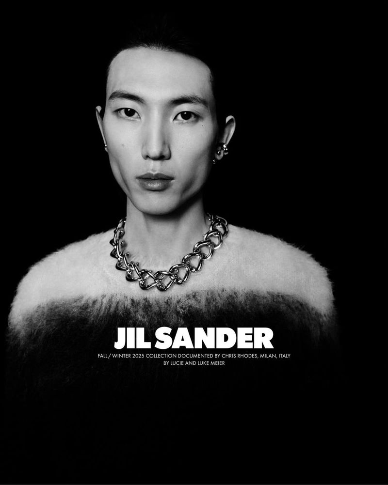 Headshot of a young adult male model with silver jewelry, in front of a dark backdrop. The model wears a textured sweater.