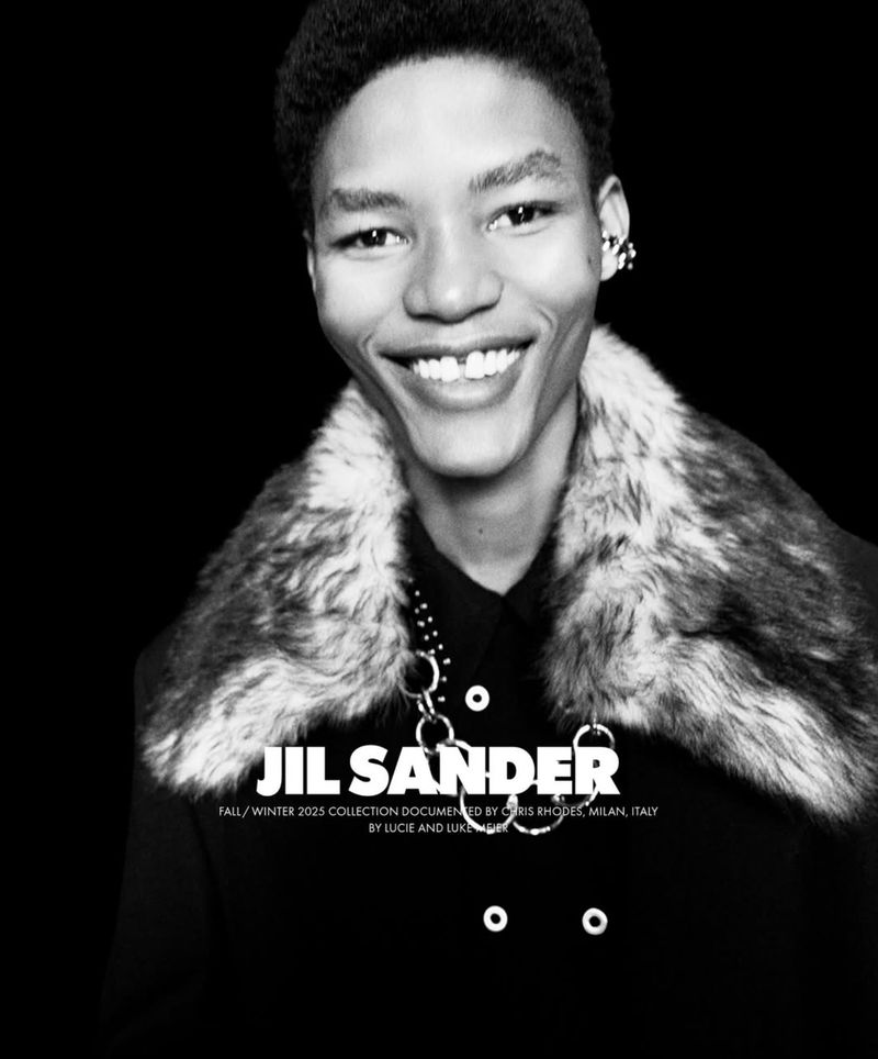 Black-and-white fashion shot of a smiling Black male model wearing a fur-collared coat and a metal-linked necklace against a dark backdrop.