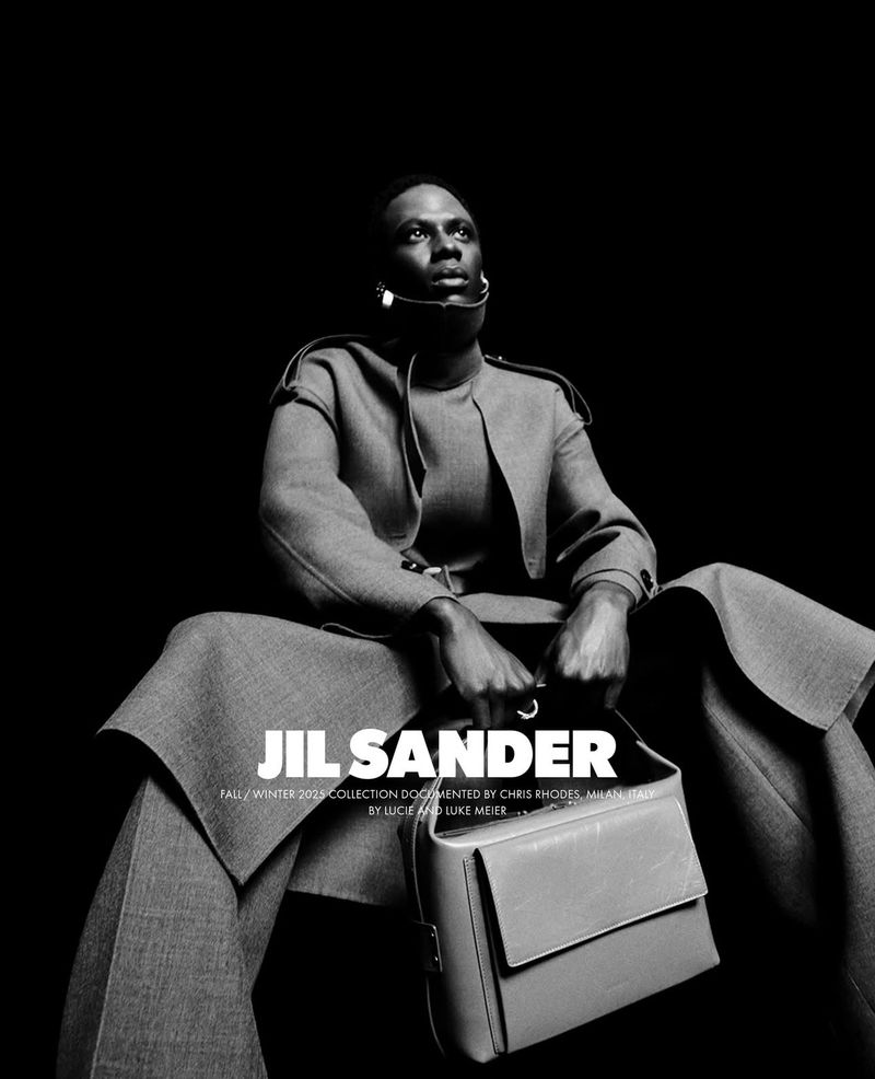 Black man seated in grey tailored coat holding a structured leather handbag against solid black