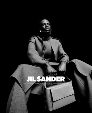 Black man seated in grey tailored coat holding a structured leather handbag against solid black