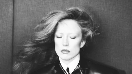 Monochrome portrait of Raquel Zimmermann with eyes closed and voluminous wind-blown hair wearing a leather jacket and white shirt.